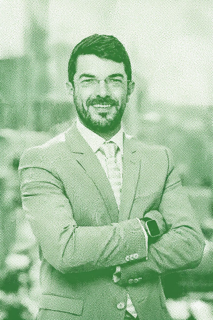 Smiling man in a suit, standing outdoors with arms crossed.